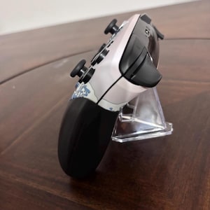 Pode incluir: Um comando de videojogos sem fios com uma placa frontal de cor clara e um padr&atilde;o de ondas azuis e brancas. O dispositivo tem joysticks pretos, bot&otilde;es superiores pretos e uma sec&ccedil;&atilde;o de ader&ecirc;ncia preta, visto de um perfil lateral.