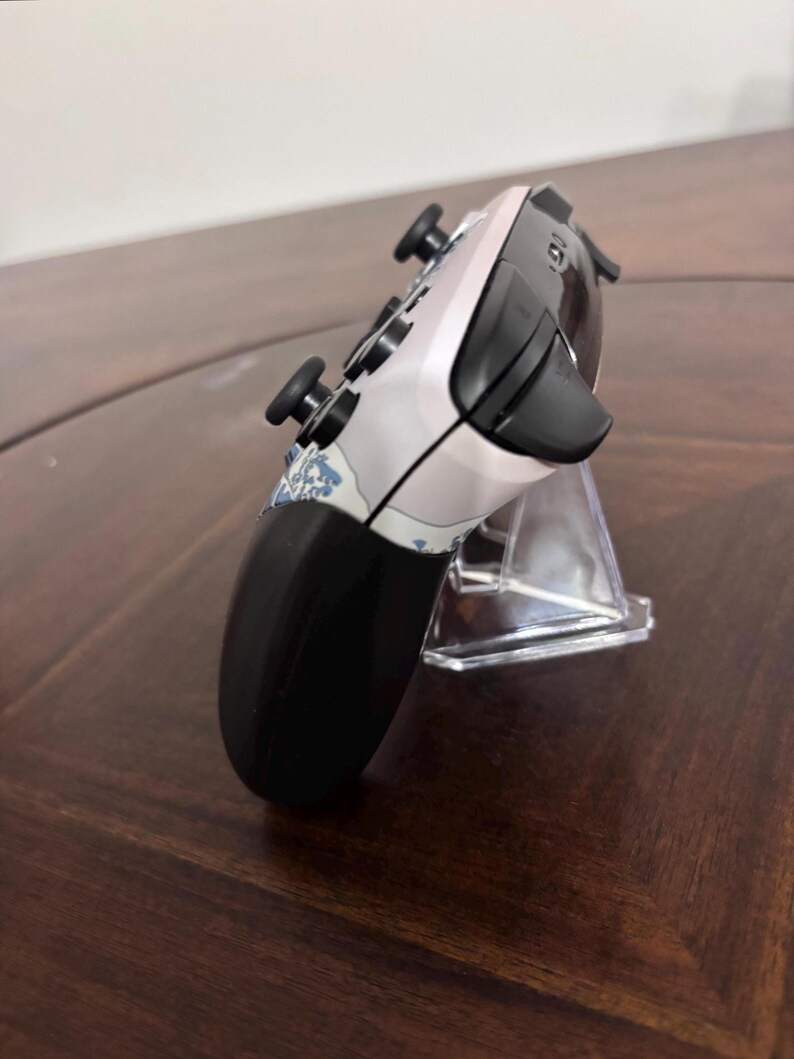 Pode incluir: Um comando de videojogos sem fios com uma placa frontal de cor clara e um padr&atilde;o de ondas azuis e brancas. O dispositivo tem joysticks pretos, bot&otilde;es superiores pretos e uma sec&ccedil;&atilde;o de ader&ecirc;ncia preta, visto de um perfil lateral.