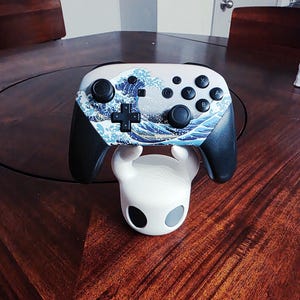 May include: A wireless video game controller featuring a Great Wave off Kanagawa graphic print on the faceplate, black grips, and black buttons, resting on a white, rounded character-themed stand with two dark circular eye-like accents.
