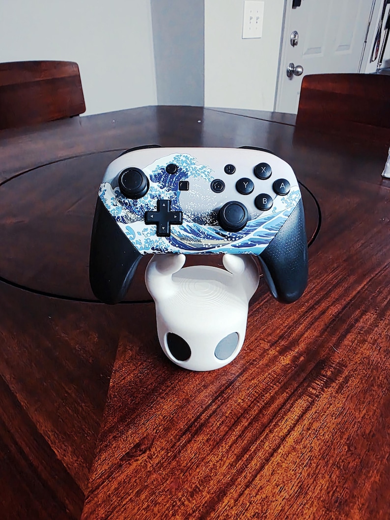 May include: A wireless video game controller featuring a Great Wave off Kanagawa graphic print on the faceplate, black grips, and black buttons, resting on a white, rounded character-themed stand with two dark circular eye-like accents.