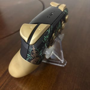 May include: A wireless video game controller featuring a black and gold color scheme with intricate green graphic patterns on the side. The device includes gold-colored shoulder buttons labeled ZL and L, a central charging port, and a clear plastic stand.