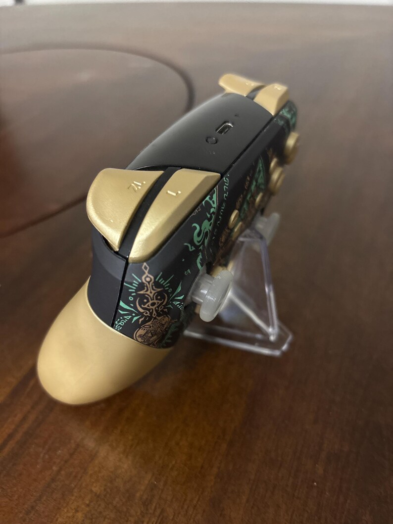 May include: A wireless video game controller featuring a black and gold color scheme with intricate green graphic patterns on the side. The device includes gold-colored shoulder buttons labeled ZL and L, a central charging port, and a clear plastic stand.