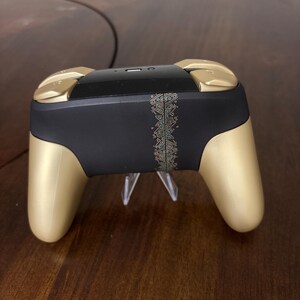 May include: A wireless video game controller featuring a matte black central body with a vertical gold-colored geometric pattern strip. The controller has gold-colored hand grips and shoulder buttons, with a glossy black top section containing a port.