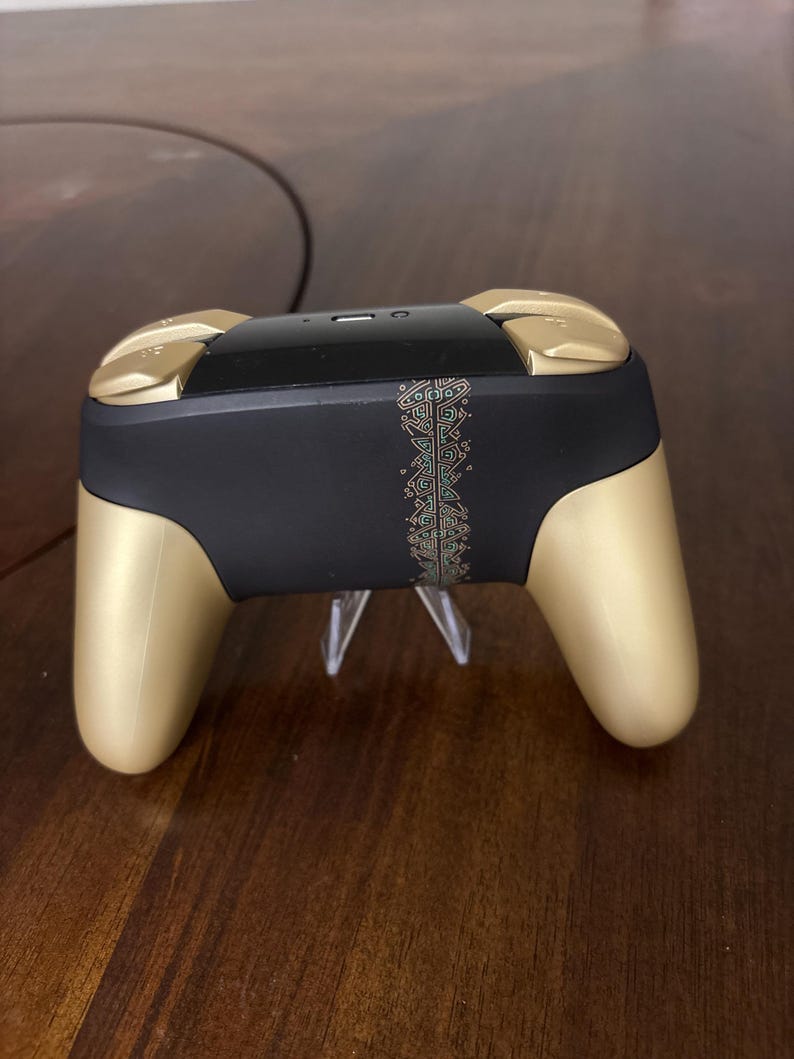 May include: A wireless video game controller featuring a matte black central body with a vertical gold-colored geometric pattern strip. The controller has gold-colored hand grips and shoulder buttons, with a glossy black top section containing a port.