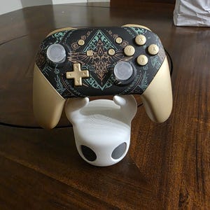 May include: A wireless video game controller with a matte gold finish and a black faceplate featuring intricate teal and gold geometric patterns. The controller rests on a white, hollow, rounded stand with two dark circular eye-like openings.
