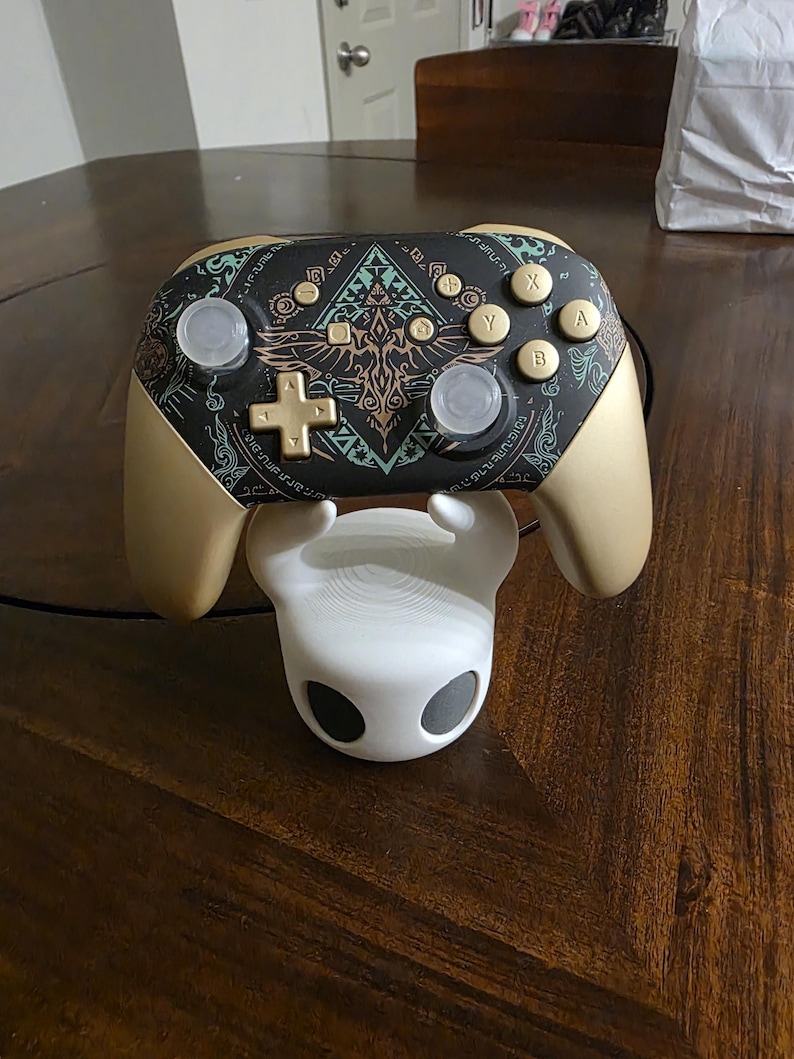 May include: A wireless video game controller with a matte gold finish and a black faceplate featuring intricate teal and gold geometric patterns. The controller rests on a white, hollow, rounded stand with two dark circular eye-like openings.