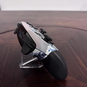 Pode incluir: Um comando de videojogos sem fios com corpo branco e cinzento, apresentando um padr&atilde;o de ondas japonesas em azul e branco. O dispositivo inclui man&iacute;pulos anal&oacute;gicos pretos, bot&otilde;es superiores pretos e uma pega preta na lateral.