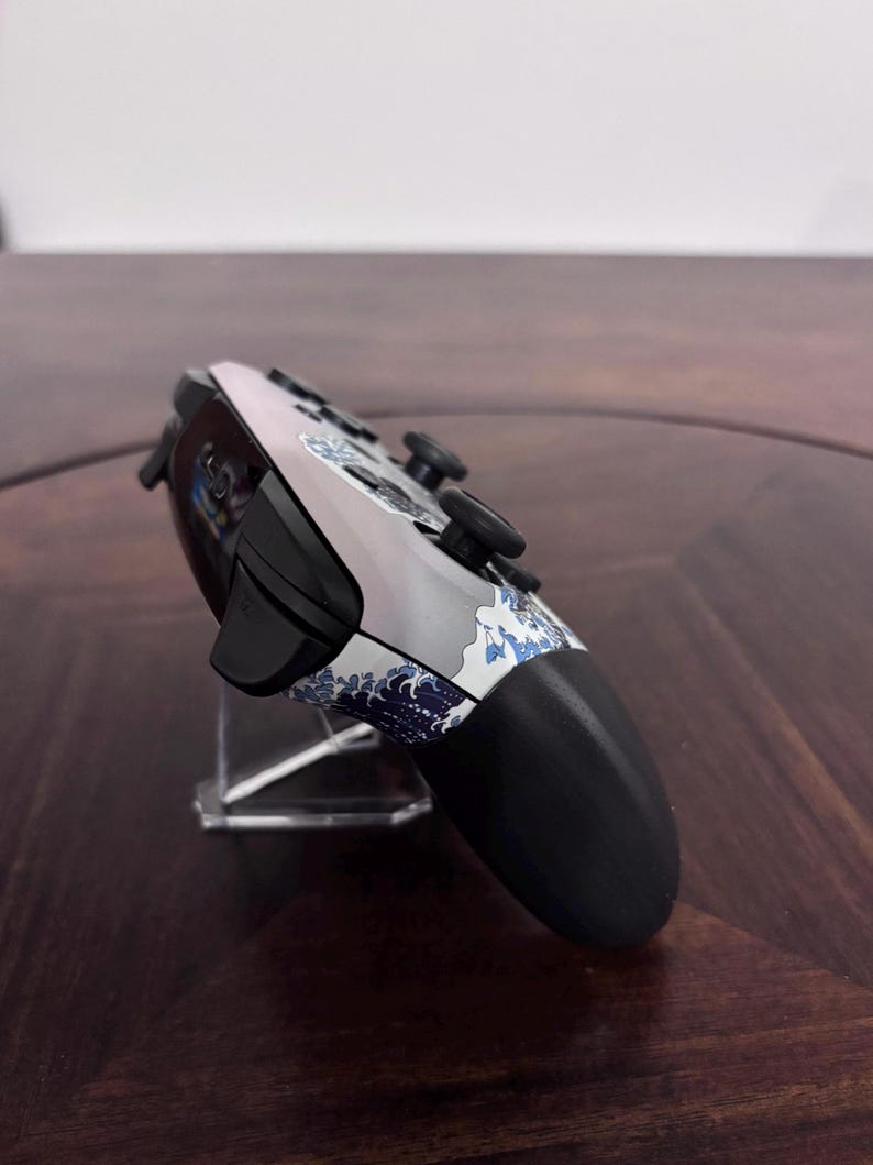 Pode incluir: Um comando de videojogos sem fios com corpo branco e cinzento, apresentando um padr&atilde;o de ondas japonesas em azul e branco. O dispositivo inclui man&iacute;pulos anal&oacute;gicos pretos, bot&otilde;es superiores pretos e uma pega preta na lateral.