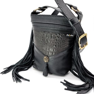 May include: Black leather crossbody bag with a fringe detail and a crocodile embossed panel. The bag has a gold buckle and a gold metal accent on the front. The bag is labeled "Fly Away".