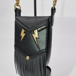 May include: Black leather crossbody bag with gold lightning bolt accents and fringe. The bag has a small pocket on the front and a strap with a gold clasp. The bag is labeled "Flip Away".