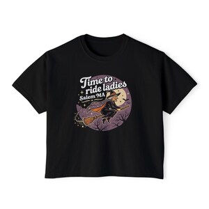 May include: A black short-sleeve t-shirt featuring a circular graphic of a witch flying on a broomstick against a full moon. The text reads Time to ride ladies and Salem MA, surrounded by stars, bats, and bare tree silhouettes in purple tones.