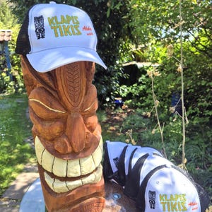 Puede incluir: Dos gorras de camionero blancas con malla negra en la parte posterior. Las gorras tienen un diseño bordado en amarillo y verde que dice "Klapís Tikis Aloha Made". Las gorras están sobre una estatua tiki de madera.