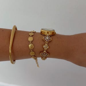 Puede incluir: Un reloj de pulsera dorado con una correa de eslabones florales con piedras transparentes, junto a una pulsera de cadena de eslabones cuadrados texturizados dorados y un brazalete r&iacute;gido dorado liso. La esfera del reloj es redonda y met&aacute;lica.