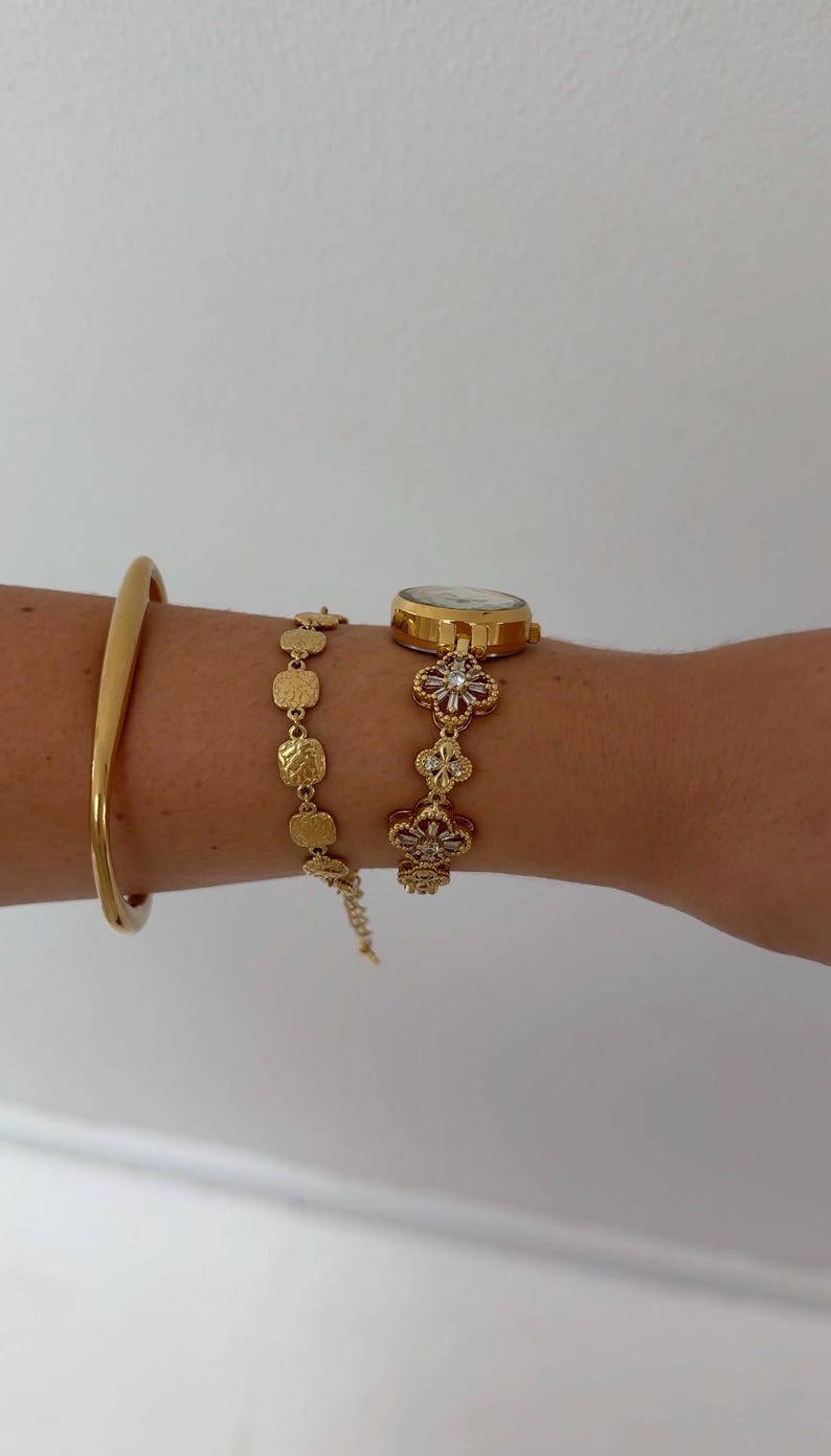 Puede incluir: Un reloj de pulsera dorado con una correa de eslabones florales con piedras transparentes, junto a una pulsera de cadena de eslabones cuadrados texturizados dorados y un brazalete r&iacute;gido dorado liso. La esfera del reloj es redonda y met&aacute;lica.