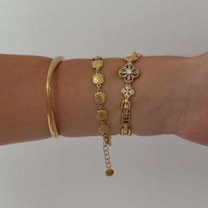 Puede incluir: Tres pulseras de tono dorado. La primera es un brazalete liso y s&oacute;lido. La segunda es una cadena delicada con peque&ntilde;os dijes circulares texturizados similares a monedas. La tercera es una pulsera de eslabones con motivos florales y piedras.