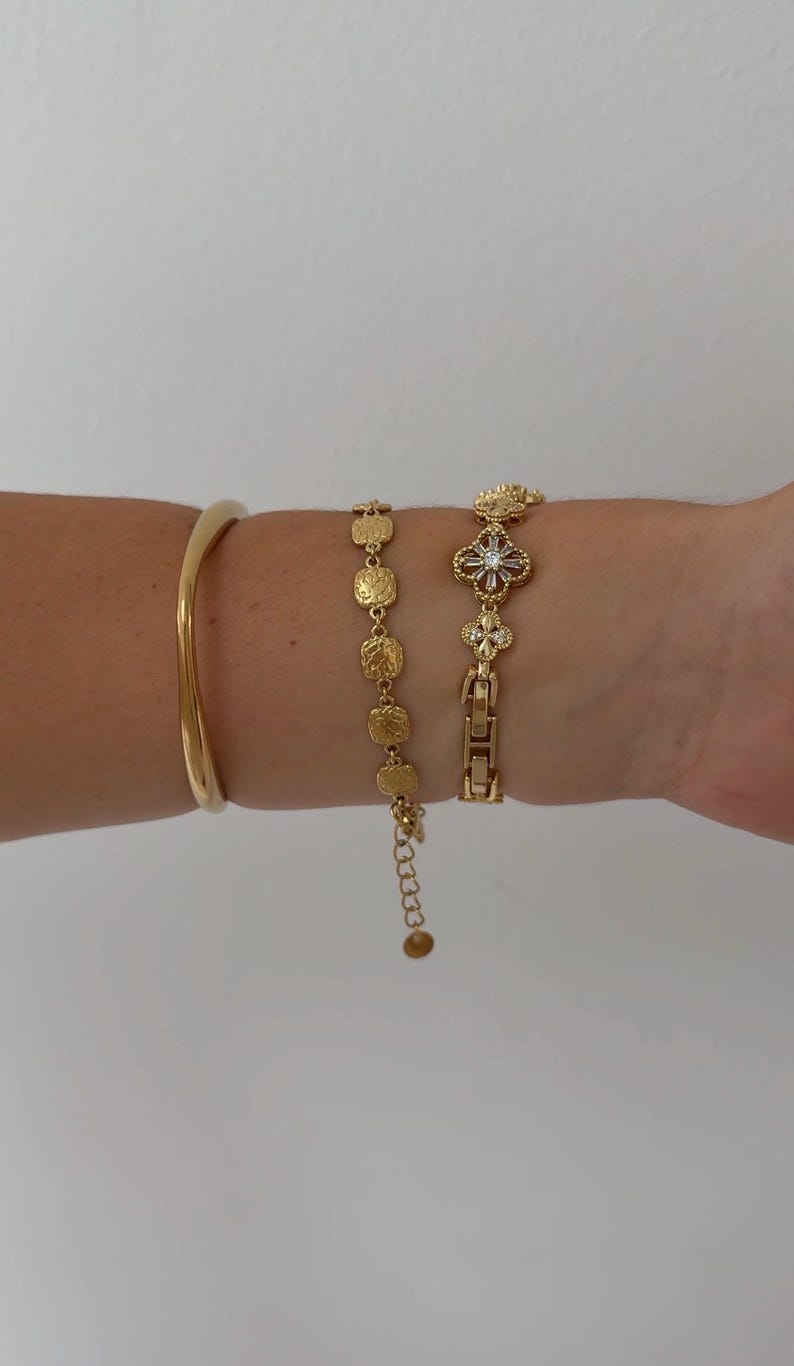 Puede incluir: Tres pulseras de tono dorado. La primera es un brazalete liso y s&oacute;lido. La segunda es una cadena delicada con peque&ntilde;os dijes circulares texturizados similares a monedas. La tercera es una pulsera de eslabones con motivos florales y piedras.