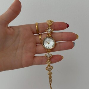 Puede incluir: Un reloj de pulsera dorado con esfera redonda blanca, manecillas e &iacute;ndices dorados. La correa est&aacute; compuesta por eslabones en forma de tr&eacute;bol de cuatro hojas, cada uno con piedras transparentes, unidos por una cadena de metal dorado.