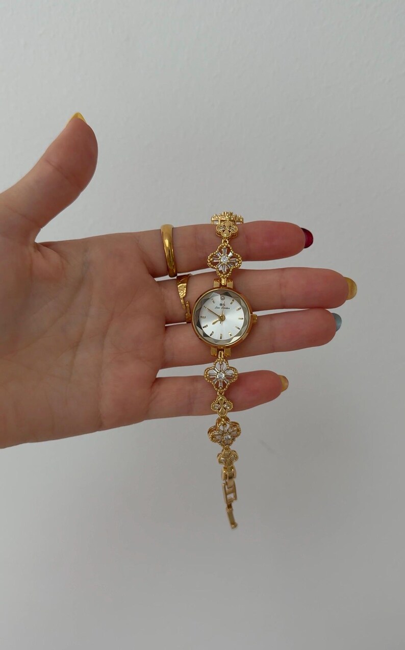 Puede incluir: Un reloj de pulsera dorado con esfera redonda blanca, manecillas e &iacute;ndices dorados. La correa est&aacute; compuesta por eslabones en forma de tr&eacute;bol de cuatro hojas, cada uno con piedras transparentes, unidos por una cadena de metal dorado.