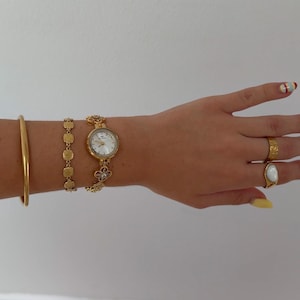 Puede incluir: Un reloj de pulsera dorado con esfera redonda blanca y eslabones en forma de flor, acompa&ntilde;ado de un brazalete fino, una pulsera de eslabones cuadrados texturizados, un anillo dorado martillado y un anillo dorado con piedra ovalada blanca.
