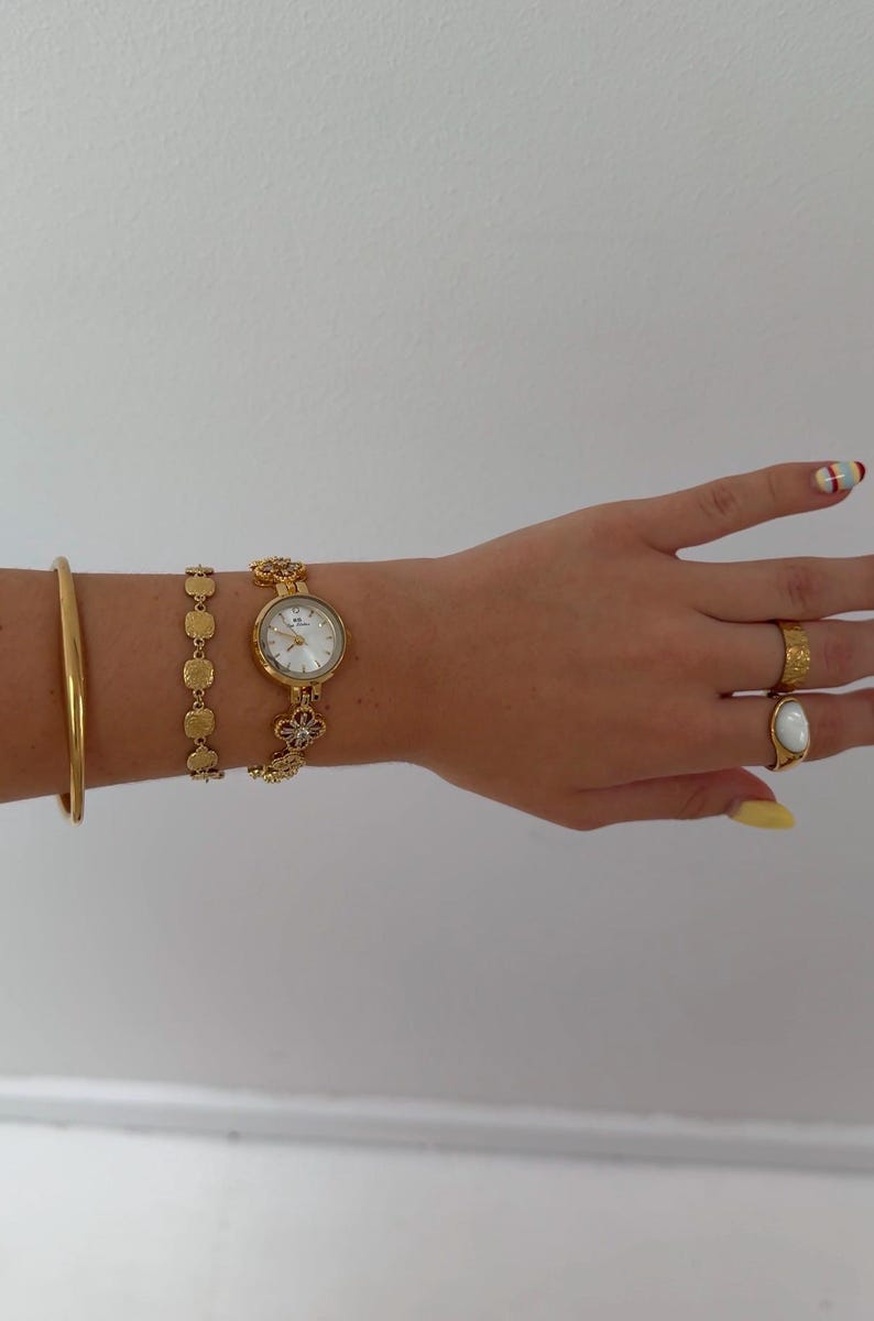 Puede incluir: Un reloj de pulsera dorado con esfera redonda blanca y eslabones en forma de flor, acompa&ntilde;ado de un brazalete fino, una pulsera de eslabones cuadrados texturizados, un anillo dorado martillado y un anillo dorado con piedra ovalada blanca.