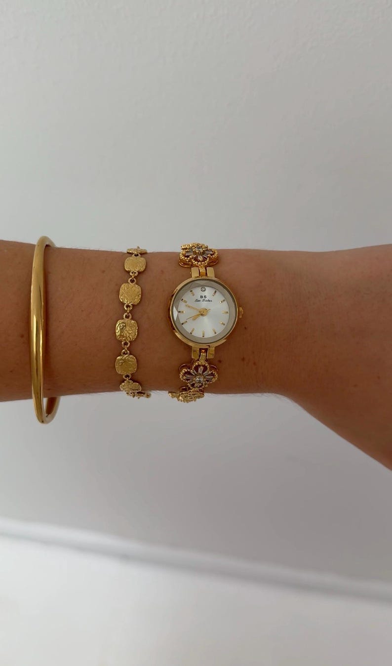 Puede incluir: Un reloj de pulsera dorado con esfera blanca redonda que muestra el texto BS Bee Sister, junto a una pulsera de eslabones florales a juego. A su lado, una pulsera de cadena con eslabones cuadrados texturizados y un brazalete dorado liso y s&oacute;lido.