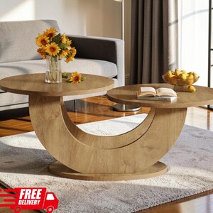 May include: A light oak wood coffee table featuring a unique crescent-shaped base that supports two circular tabletops at different heights. The table holds a clear glass vase filled with yellow sunflowers and a bowl of yellow pears with a book.