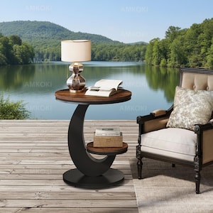 May include: A round side table with a dark wood top and a black C-shaped base featuring a lower shelf. A table lamp with a white shade and a glass base sits on the top surface next to an open book. A small wooden box rests on the lower shelf.