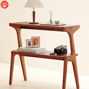 Op de afbeelding: Een houten console met twee niveaus, met een mid-century modern esthetiek. De warme houten tafel bevat een lamp, ingelijste foto's, boeken, een camera en decoratieve items. De kenmerkende vorm en het ontwerp van de tafel maken het een stijlvolle toevoeging aan elke woonruimte.