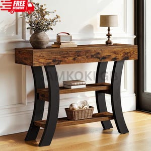 May include: A console table with a dark wood grain top and two lower shelves supported by four curved black metal legs. The table holds a vase with dried branches, a small rectangular box, and a lamp. A basket and books sit on the shelves.