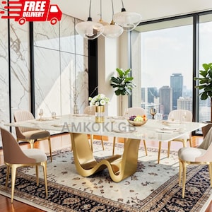 May include: A rectangular dining table with a white marble-patterned top and a gold-colored, wave-shaped pedestal base. The table is surrounded by six upholstered chairs with gold-colored metal legs, set on a patterned area rug in a room.