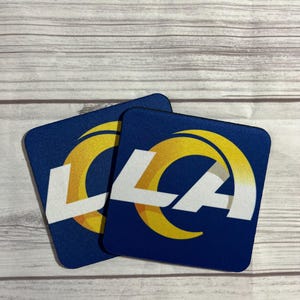 May include: Two blue and yellow coasters with the Los Angeles Rams logo. The logo is a white "LA" with a yellow crescent moon behind it.