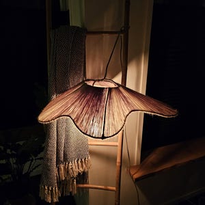 Puede incluir: Una pantalla de lámpara tejida con una forma orgánica única cuelga de una escalera de madera. La pantalla está encendida, proyectando un cálido resplandor en el área circundante.