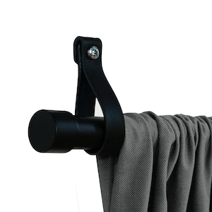 Peut inclure: Tringle à rideau en métal noir avec un embout en cuir noir. La tringle est fixée au mur et un rideau gris est suspendu à celle-ci.