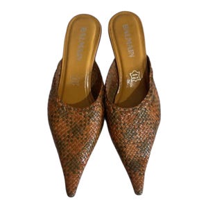 May include: A pair of brown and orange patterned slip-on mules with pointed toes. The shoes have a woven texture and a leather insole with the brand name "BALMAIN" printed in gold. The heels are low.