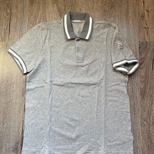 Puede incluir: Un polo gris con cuello y puños a rayas blancas y grises. La camisa tiene una tapeta de tres botones y un parche con el logo en la manga izquierda. La tela es suave y transpirable.