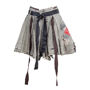 May include: A grey patchwork skirt with a flared shape. The skirt has vertical stripes, dark ribbons, and a red patch. Brown ribbons hang from the hem. The skirt has a button closure and a black ribbon tie at the waist.