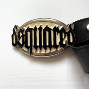 May include: A silver-toned belt buckle with the word "DSQUARED" in black gothic lettering. The buckle has a cream-colored background and is attached to a black leather belt.