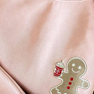May include: A light pink sweatshirt with a gingerbread man design. The gingerbread man is holding a red and white cup with whipped cream. The gingerbread man has a smiling face and red buttons.