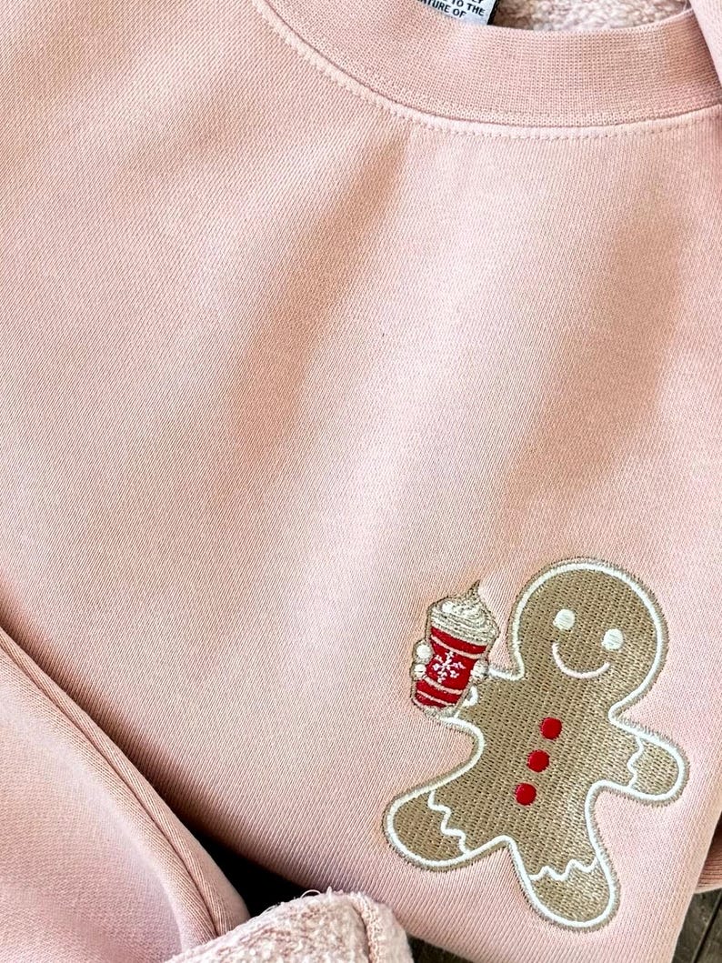 May include: A light pink sweatshirt with a gingerbread man design. The gingerbread man is holding a red and white cup with whipped cream. The gingerbread man has a smiling face and red buttons.
