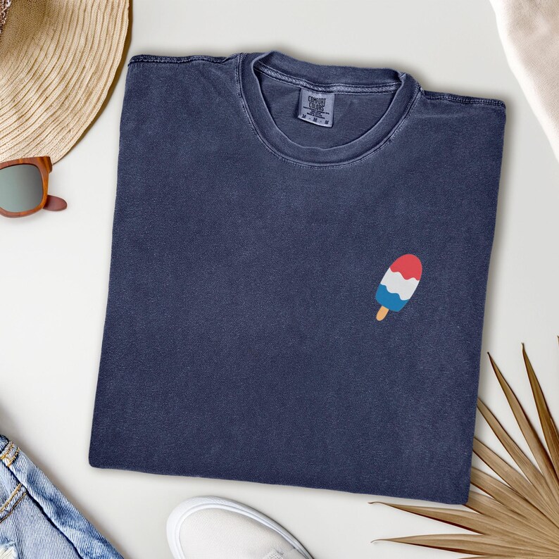 May include: Navy blue t-shirt with a small embroidered popsicle design in red, white, and blue. The shirt is made of a soft, comfortable material, perfect for casual wear. The shirt is folded neatly on a white surface.