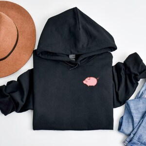 K&ouml;nnte beinhalten: Schwarzer Kapuzenpullover mit einer rosa Schwein-Illustration auf der Vorderseite.