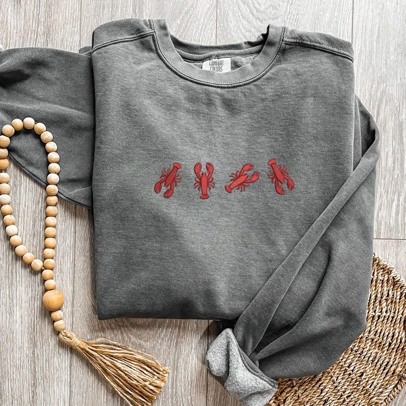 May include: A charcoal gray sweatshirt with four embroidered red lobsters across the chest. The sweatshirt is folded, with the sleeves and hem visible. A wooden bead garland and woven basket are in the background.