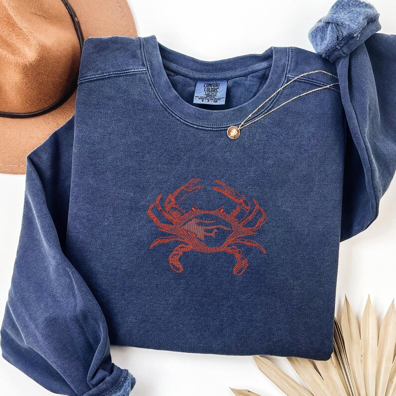 K&ouml;nnte beinhalten: Ein marineblaues Sweatshirt mit einem roten Krebsdesign. Das Sweatshirt hat einen Rundhalsausschnitt und lange &Auml;rmel. Eine goldene Halskette mit Anh&auml;nger ist &uuml;ber dem Sweatshirt drapiert. Ein brauner Hut ist im Hintergrund.