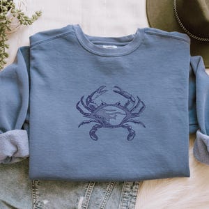K&ouml;nnte beinhalten: Ein blaues Sweatshirt mit Rundhalsausschnitt und langen &Auml;rmeln. Eine detaillierte marineblaue Krabben-Grafik ist mittig auf der Vorderseite. Das Sweatshirt ist auf einer Jeans gefaltet.