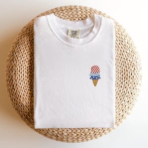May include: White t-shirt with a small embroidered ice cream cone design. The ice cream cone features red and white stripes, blue stars, and a brown cone. The shirt is folded on a woven, circular surface.