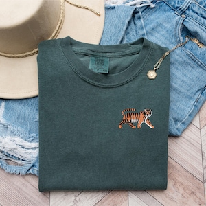 May include: A dark green t-shirt with a tiger embroidered on the front. The tiger is orange and black with white stripes. The t-shirt is folded and laid on a denim background.