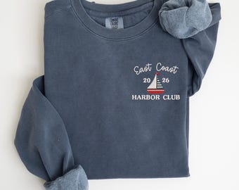 May include: A blue-grey sweatshirt with the text "East Coast Harbor Club" and a sailboat graphic. The sweatshirt has a crew neck and long sleeves with cuffed wrists. The Comfort Colors tag is visible.