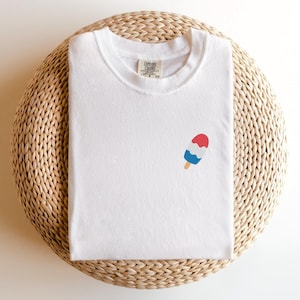 May include: White t-shirt with a small popsicle graphic in red, white, and blue. The shirt is folded and placed on a woven, circular surface. The tag reads "Comfort Colors".