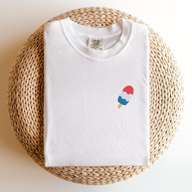 May include: White t-shirt with a small popsicle graphic in red, white, and blue. The shirt is folded and placed on a woven, circular surface. The tag reads "Comfort Colors".