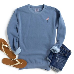 May include: A blue crew neck sweatshirt with a small pink flamingo embroidered on the chest. The sweatshirt is paired with denim shorts and brown sandals with gold straps, all on a white background.